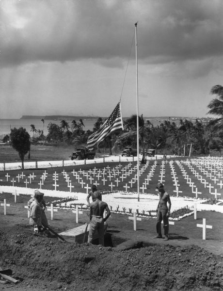 The cemetery for the war dead on Guam. The body of a fighting man who died of wounds received during the battle for Iwo Jima is being lowered into the earth. In the background trees are along a shoreline, and a far shoreline is in the distance.