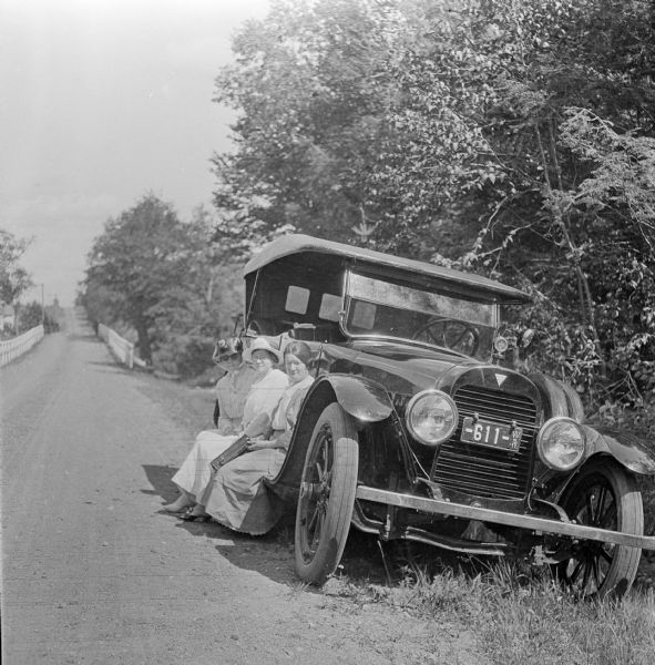 Three women sitting on the side rail of a Hudson Touring Car along a country road.