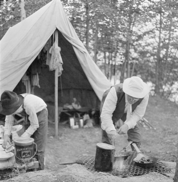 Franklin E. Bump, brother-in-law of Dr. Smith, and one of his children cooking on a fire pit in front of their campsite. The campsite is situated in the woods next to a lake.