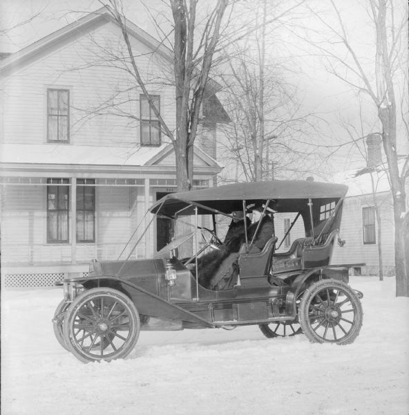 Winter scene with Dr. Joseph Smith and his wife, Mary E. Smith, sitting in an automobile parked in front of a house.  Snow is on the ground and the automobile has chains attached to the back tires.