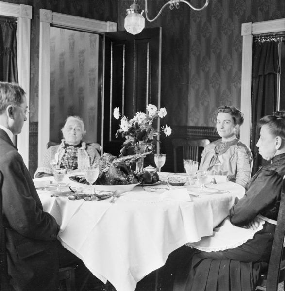 Dr. Joseph Smith and Mary E. Smith seated at a set dinner table with Harriet Millard and Harriet Millard Smith. On the table is a bouquet of flowers and a serving dish of roasted poultry.