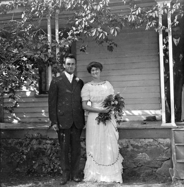 Wedding day of Alice Armstrong Ashmun, Mary's niece, to Edwin Trenbath.  They are standing in wedding attire in front of a porch while Alice is cradling a bouquet of flowers in her arms. Other family members can be seen standing in the doorway of the house.