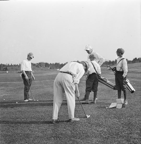 Dr. Joseph Smith golfing at Wausau Country Club with friends. One man is putting while two other men stand by watching. Two young caddies carrying clubs stand amongst the men.
