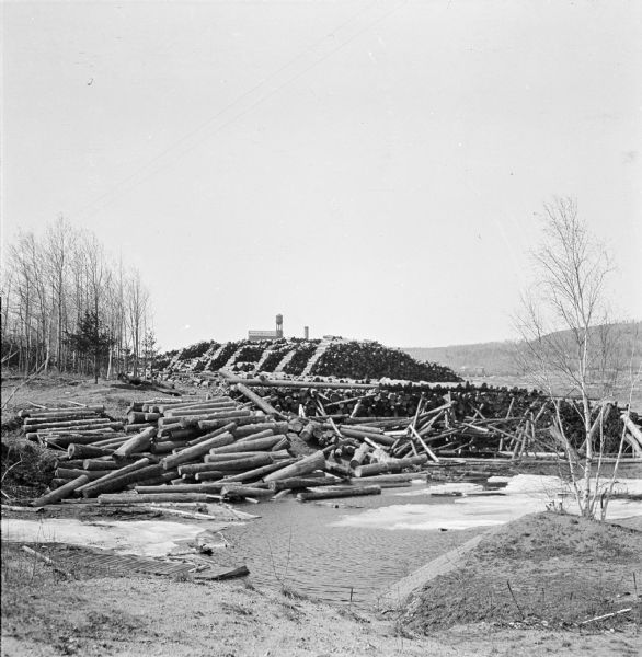 View from low hill of piles of logs along the icy rivers edge. A pile of logs is on the shoreline, and a large amount of neatly stacked logs are in the background. Far behind the stacked logs is a water tower, smokestack and the top of a large building.