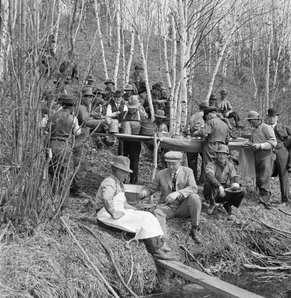 View towards shoreline of log drivers eating lunch on the edge of the riverbank. Visiting businessmen share a meal with the log drivers who are sitting and eating on the steep bank. A table is set up buffet style with provisions, and a man wearing an apron sits at the river's edge near one of the businessmen.