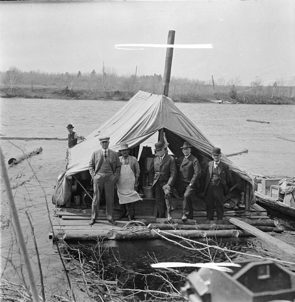 View looking down from riverbank of four businessmen and the log driving cook posing on the cooks tent barge during the log drive.