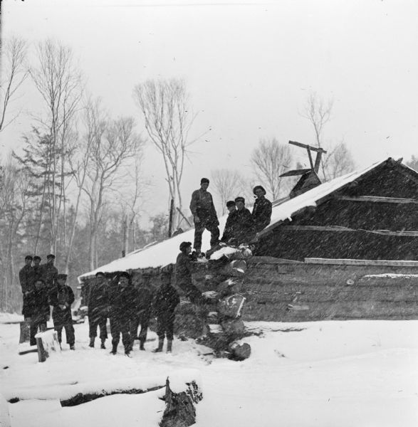 Several loggers are standing next to a log cabin, while a few men are sitting on the roof of the cabin during a snowstorm.