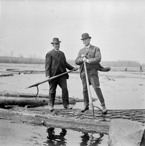 Two businessmen visiting a log drive along the Wisconsin River.  They stand atop a log holding peavey hooks.