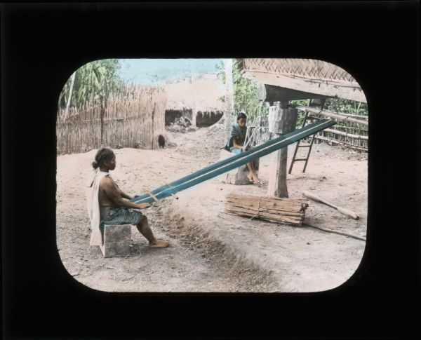 Outdoor view of one woman using a loom and another woman preparing fibers for the loom to create siding for housing. This image was taken or purchased by Carrie Chapman Catt during her trip to Ceylon (now Sri Lanka). In her journal from Ceylon she records: "The women braid the fibres together making a crude mat of them. The trunks of the trees are set up in the form of a house and these mats are hung up to make the walls."