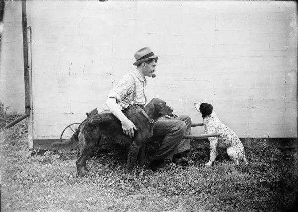 Outdoor portrait of Kent Wood, with a pipe in his mouth and sitting in a cart, posing with two dogs. All three are in profile. The side of a building is behind the group.