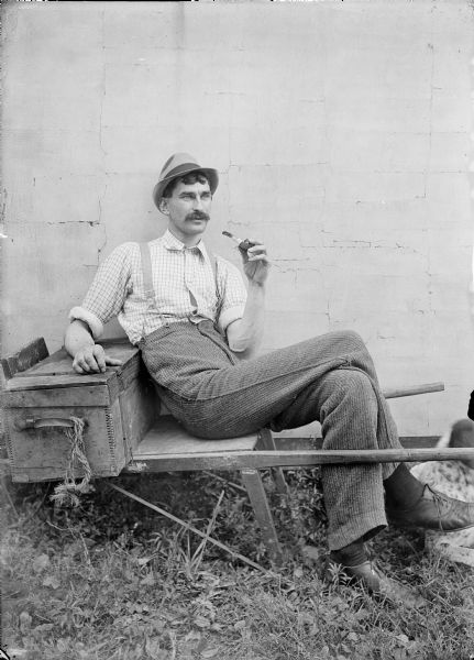 Outdoor portrait of Kent Wood sitting on a wooden wheelbarrow against a wall holding a pipe. He has his right arm resting on a wooden box. A dog (at the edge of the image at right) is lying at his feet.