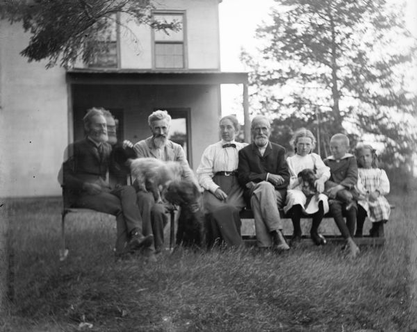 Outdoor group portrait of the Thwaites-Turvill family. Seven members of the family, and three dogs, are sitting on chairs and a bench in the front yard of their family home.