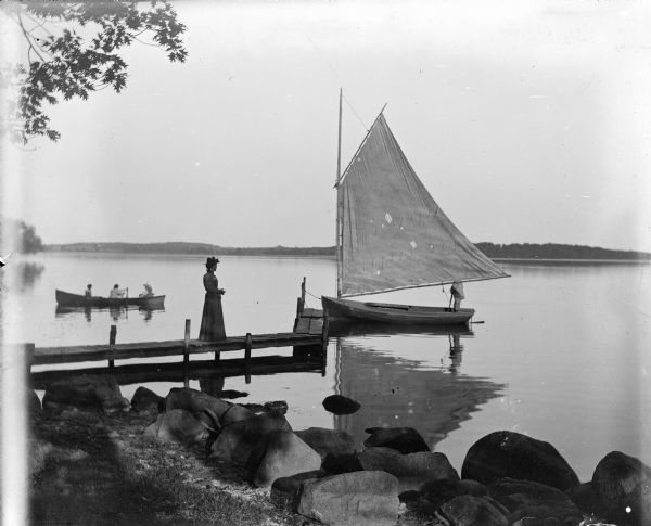 View from rocky shoreline of Turvill's new sailboat tied to a dock. A women is standing on the narrow wooden dock. A man is standing in the sailboat, with his face blocked by the full sail. On the left behind the dock are three people in a rowboat. The far shoreline is in the background.