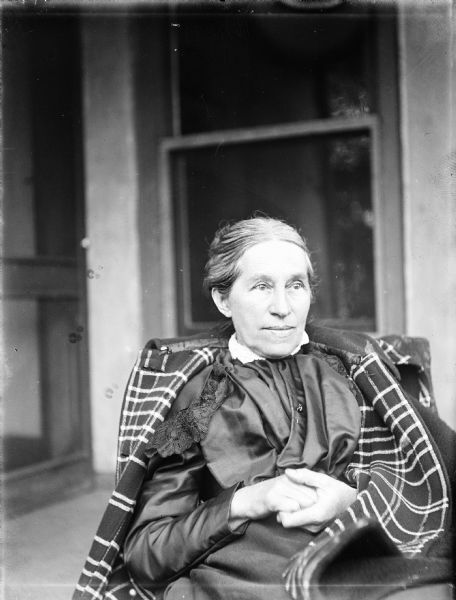 Outdoor portrait of Elizabeth Turvill Wood. She sitting in a chair in front of a window, with a coat draped over her shoulders and her hands clasped in her lap.