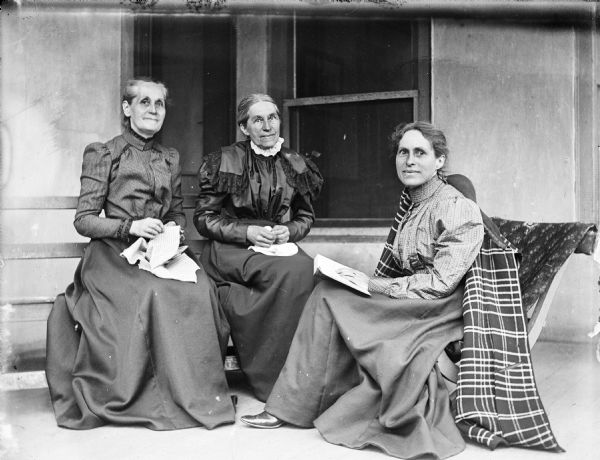 Outdoor family portrait of Mary Turvill McConnell, Elizabeth Turvill Wood, and Jessie Turvill Thwaites. They are sitting on a porch in front of windows with either hand sewing or a book in their laps. Jessie Thwaites has a coat draped over her right shoulder.