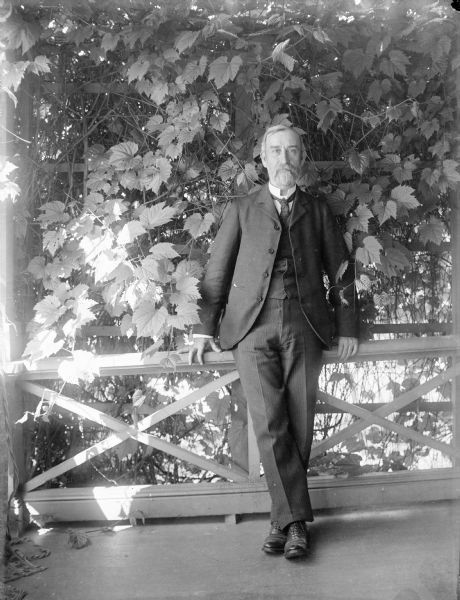 Outdoor portrait of Thomas Turvill standing on a porch. He is seen leaning against the railing and trellis which is covered in foliage.