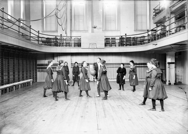 University of Wisconsin-Madison freshman women's basketball team in the gymnasium of Ladies Hall (now Chadbourne Hall). A man stands in the center holding a basketball for the tip-off.