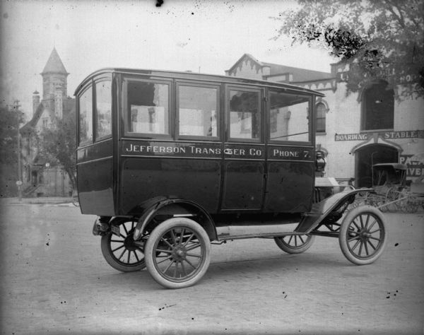 A bus for the Jefferson Transfer Company sits in front of the Schoelkopf Automobile dealership on East Washington Avenue. Behind the bus is a Boarding Stable and the Palace Livery. The bus is a 1915 Model T. 