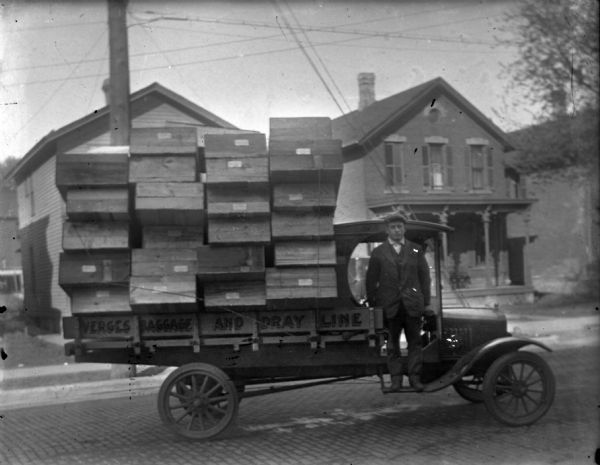 Verges Baggage and Dray Line Passenger side view of a Verges Baggage and Dray Line truck No. 5. The back of the truck is loaded with stacks of boxes. The driver of the truck is standing on the running board. Probably 134 (left) and 132 (right) S. Franklin St.