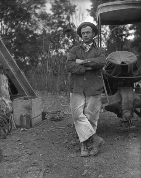 Man Standing in Outdoor Work Area Man standing near a large piece of machinery in an outdoor work area. He is wearing dirty canvas pants, work boots and a hat.