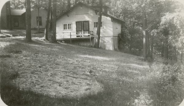 A simple wood frame cottage is built into the slope of the hill above Geneva Lake at Holiday Home Camp. A second building is seen in the background.