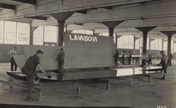 Several men paint and prepare wing assembly sections for a Lawson Air Liner. In the background more men are working at a table. Alfred Lawson is standing to the right of them.