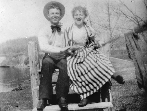 Young Man and Woman on a Stile A laughing young man and woman sitting on a stile. The man is wearing a straw hat and bow tie; she is wearing a striped dress and matching jacket. The woman has one of her feet up in the air and is also holding a stick (blurred movement) with what appears to be a jacket hung over it.