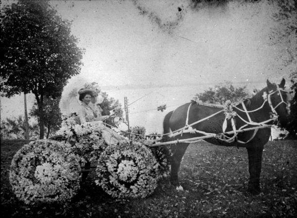 Two Women in a Flower Bedecked Horse-Drawn Cart Two women in fancy hats and summer dresses are sitting in a horse-drawn cart which has flower bedecked wheels and seat. One of the woman has an umbrella. In the background below them is a lake and far shoreline.
