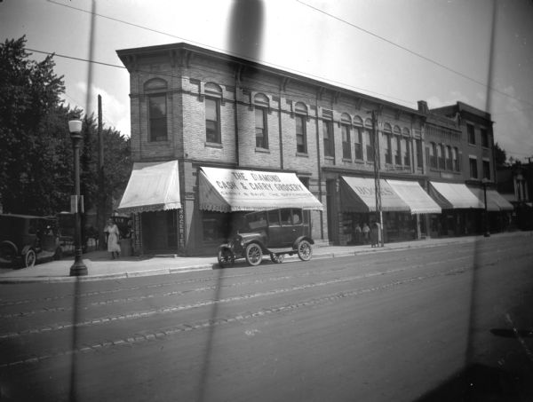 Diamond Cash and Carry Grocery View from across street towards the Diamond Cash and Carry Grocery at 334 State Street at the corner of East Gorham Street. Next door on the right is the Brown Book Shop. There is a woman in a hat and light-colored summer dress walking near the grocery. An automobile is parked on State Street. On the street around the corner is a horse-drawn vehicle parked between two cars.