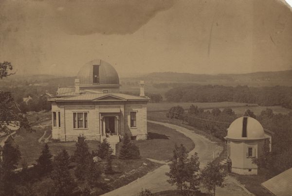 Elevated view of the University of Wisconsin-Madison Washburn Observatory. Hill, trees and fields are in the background. A man is standing near a small observatory on the right. A man and another person are posing on the steps of the Washburn Observtory.