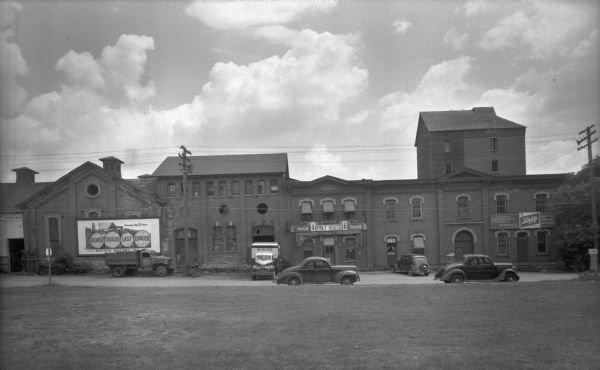 View across lawn towards the Malt House at 1603 Sherman Avenue. To the right of the Malt House is the Cafferty Distributing Company, wholesalers for Schlitz beer — "Just the Kiss of the Hops." On the left is a large billboard for Kayser Motors Inc., with the slogan: "Ford Trucks Last Longer." A Cafferty delivery truck is parked below the billboard. Directly next to the Malt House is a Schlitz truck. Three automobiles are parked along the curbs. There is a sign for Lake Mendota on the far right. 