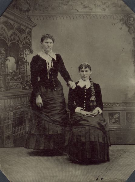 Full-length ferrotype/tintype of Martha and Esther Lomas from Lancaster, Wisconsin, in front of a painted backdrop. One of the women is standing with her left hand on the right shoulder of a woman who is sitting in a chair with a book in her lap. The seated woman has a long braid hanging down her left shoulder. Hand-coloring on cheeks and gold details on ring of woman standing, and bracelet of woman sitting.
