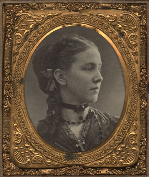 Cased albumen print of Irene Larkin, a school teacher. Bust figure facing right, wearing a choker, necklace, and a cross at her lace collar. She has a bow behind her right ear. 