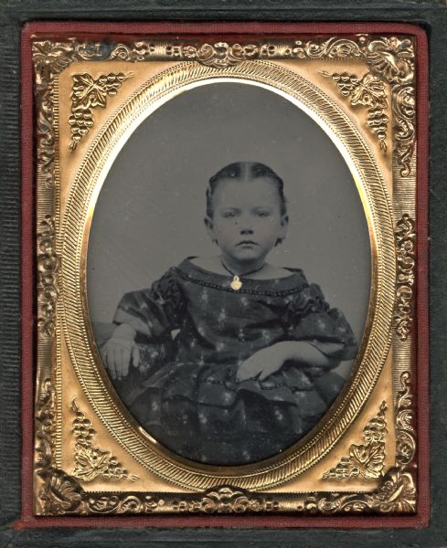 Nancy Judson Sanford Ninth plate ferrotype/tintype of Nancy Judson Sanford as a child. Three-quarter length portrait, seated, facing front, with her arm resting on a cloth-covered table, the other in her lap. She is wearing an off-the-shoulder printed dress with full sleeves and ruffled skirt, and a choker with gold pendant. Hand-coloring on cheeks and pendant,