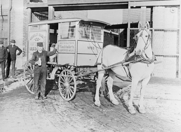 A man holding a bottle of milk is standing and posing next to a horse-drawn Gridley Dairy milk wagon. Two other men are standing on the left near a brick building in the background.
