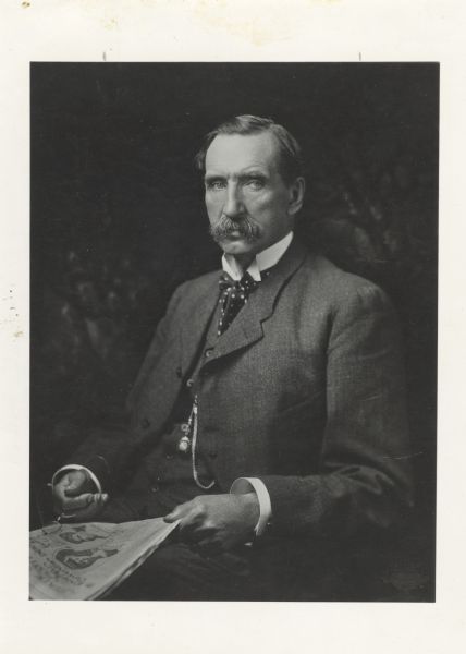Portrait of Governor William Dempster Hoard Waist-up seated portrait of Wisconsin's 16th governor, William Dempster Hoard. He is wearing a suit, a polka dot bow tie, and a watch fob on his vest. He is holding a newspaper in his left hand and eyeglasses in his right hand.