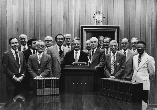 Governor Lucey Receiving the Encyclopedia Britannica Patrick J. Lucey standing at a podium (which is sitting on a table) in the State Historical Society surrounded by a group of men. Behind the group on the paneled wall is a portrait painting and a clock. The encyclopedia Britannica is displayed on the table to the left and right of the podium.