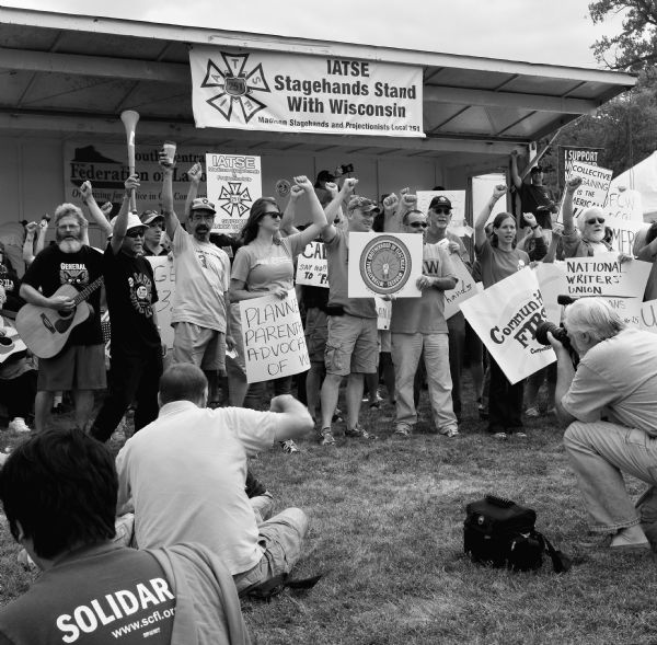 A crowd of men and women standing with fists raised in the air and holding signs at the Union Temple on Labor Day. A number of people in the foreground are sitting on the grass, and one man is crouching holding a camera. The signs that people are holding are for various unions and other social issues, such as Planned Parenthood, the National Writers' Union, and the International Brotherhood of Electrical Workers. A large sign hanging above the crowed on the awning reads: "IATSE Stagehands Stand with Wisconsin: Madison Stagehands and Projectionists Local 251."