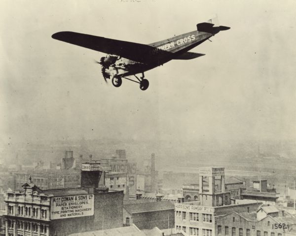 Fokker F-VII Triomotor Aerial view of a Fokker F-VII Triomotor in flight flying low over a buildings in a city. This airplane, called the "Southern Cross," was flown in the spring of 1928 from the United States to Australia in the first trans-Pacific flight. The crew included Captain Charles Kingsford Smith, Flight Lieut. Charles Ulm, navigator Harry Lyon, and radio operator James Warner. Advertisements seen on the buildings below read: "Alex Cowan & Sons Ltd. Paper envelopes, stationery, type, printing machinery and materials," "Cowan + Sons Ltd.," "Yes Bros.," "Harrisons Ramsay Pty. Ltd."