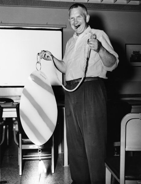 Portrait of Sid standing in a room holding an oversized fishing lure attached to a rope. Sid has a cigar in his mouth, and his fingernails are stained, probably with pyrogallol, a staining agent used in the development of film. Behind Sid are tables and chairs, and a screen on the back wall.
