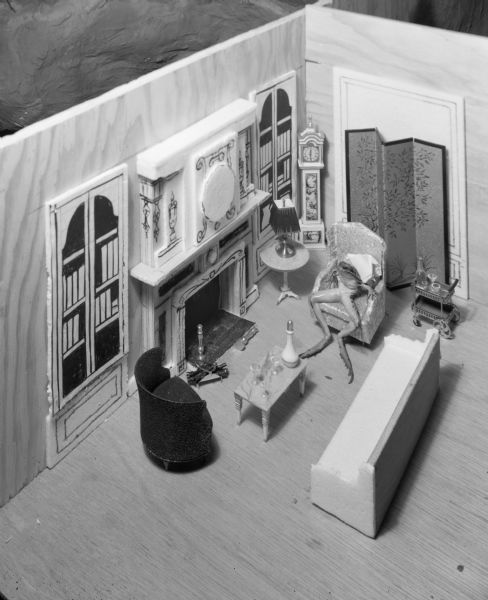 View looking down at a diorama of a living room with doll-sized furniture. A frog is sitting in a wing-back chair next to a fireplace.