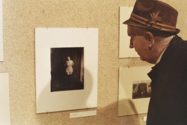 Sid Looking at Photography Exhibit Sid looking at a "Storyville" photograph by E. J. Bellocq at an exhibition. He is wearing a brown fedora with buckled band and feather.