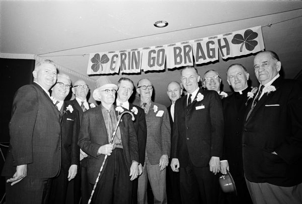 Group portrait of eleven men at Joseph "Roundy" Coughlin's Saint Patrick's Day party. Behind them is a sign with the words "Erin Go Bragh." During the party there was a singing contest between men of Irish and Norwegian descent, with the Irish prevailing. "Roundy," sixth from the left, is shown with a few Irishmen and a group of Norsemen.