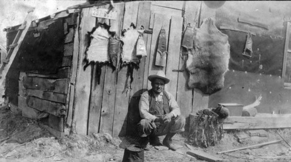 Trapper with Hides A Native American trapper sits on his haunches in front of a building where various hides on stretchers have been hung to dry. The building is sided with slab lumber and tarpaper. A ladder leads to the roof, and a license plate hangs on the front wall. There are a number of traps with chains attached on the right.