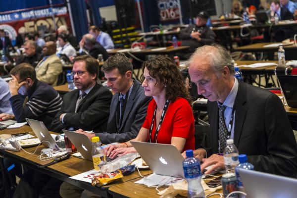 "Milwaukee Journal Sentinel" journalists sitting at tables. From left to right: Rick Wood, Dan Bice, Patrick Marley, Mary Spicuzza, and Craig Gilbert are working in the media filing center at the Republican presidential debate. Spicuzza, Gilbert, and Marley are typing on their computers, while Bice is sitting back with his arms folded watching the debates. Wood is leaning forward resting his head in his hand while looking down at his computer.