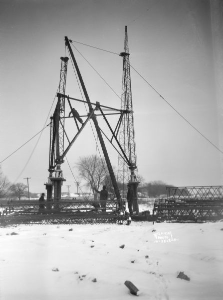 View across snow-covered ground towards four men constructing the W.I.B.A. radio transmission tower.