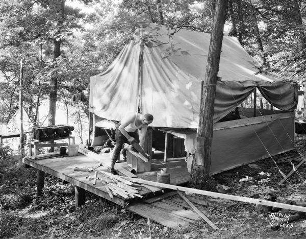 Man sawing wood in front of a tent in the woods at the University of Wisconsin Tent Colony. The tent is set up on a wood platform. Also known as Camp Gallistella on the south shore of Lake Mendota, west of second point.
