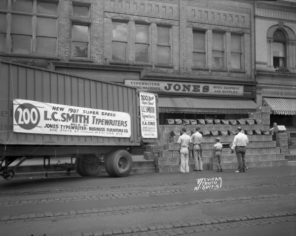 L.C. Smith Typewriter Truck View across street towards a semi-trailer truck delivering 200 new 1937 Super Speed L.C. Smith typewriters. A young boy and three men are standing in the street looking at L.C. Smith typewriter boxes stacked up in front of the Jones Typewriter — Business Furniture Co., 506 State Street.