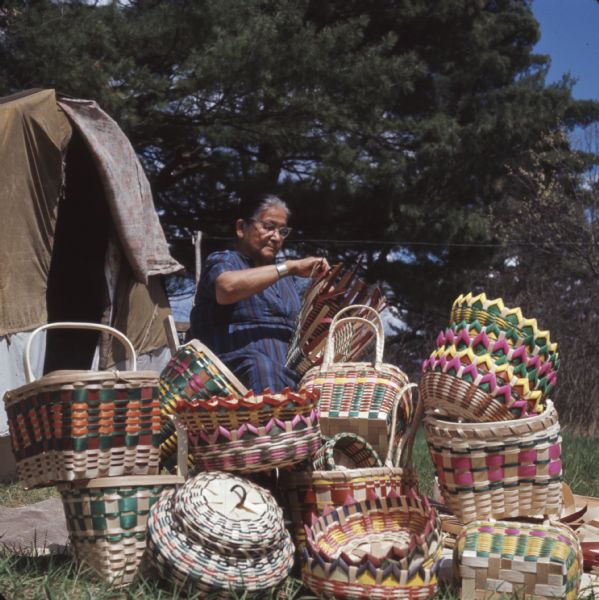 A Ho-Chunk woman, Mable Davis (maiden name) Lowe is sitting and weaving a basket. Behind her is a dwelling (chipoteke). Several finished woven baskets are displayed in front of her.