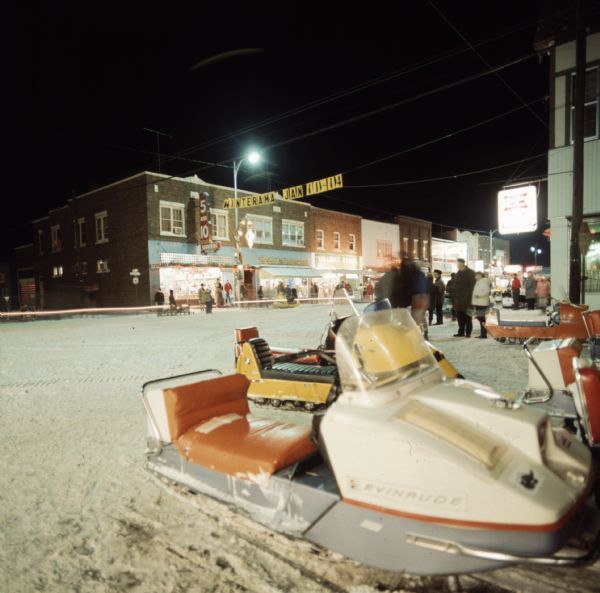 Groups of people are walking or standing downtown along the snow-covered street at night. Snowmobiles are parked in the right foreground. A banner stretching between the buildings on either side of the street reads: "Winterama Jan 11-14."